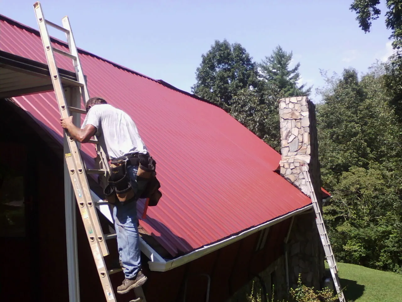 Roofer on a ladder beside a red metal roof and stone chimney — Soffit & Fascia Repair by Roofing Brothers in Packwood